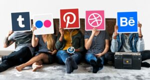 Five people sitting on a couch each holding up a large icon of different social media platforms covering their faces. Social media icons include Tumblr, Flickr, Pinterest, Dribbble, and Behance.